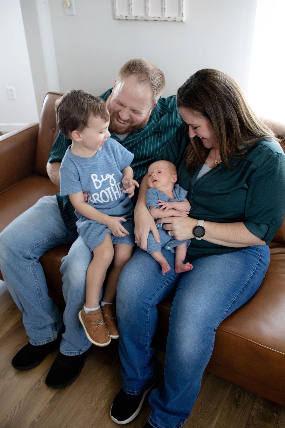 Family of four sits on leather sofa, parents smiling at newborn baby while toddler in Big Brother shirt looks on.