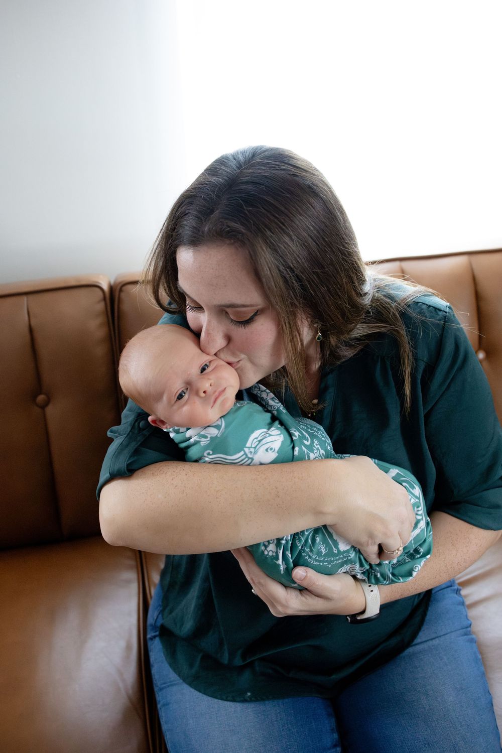 A mother tenderly kisses her newborn baby wrapped in a teal swaddle while sitting on a brown leather sofa.
