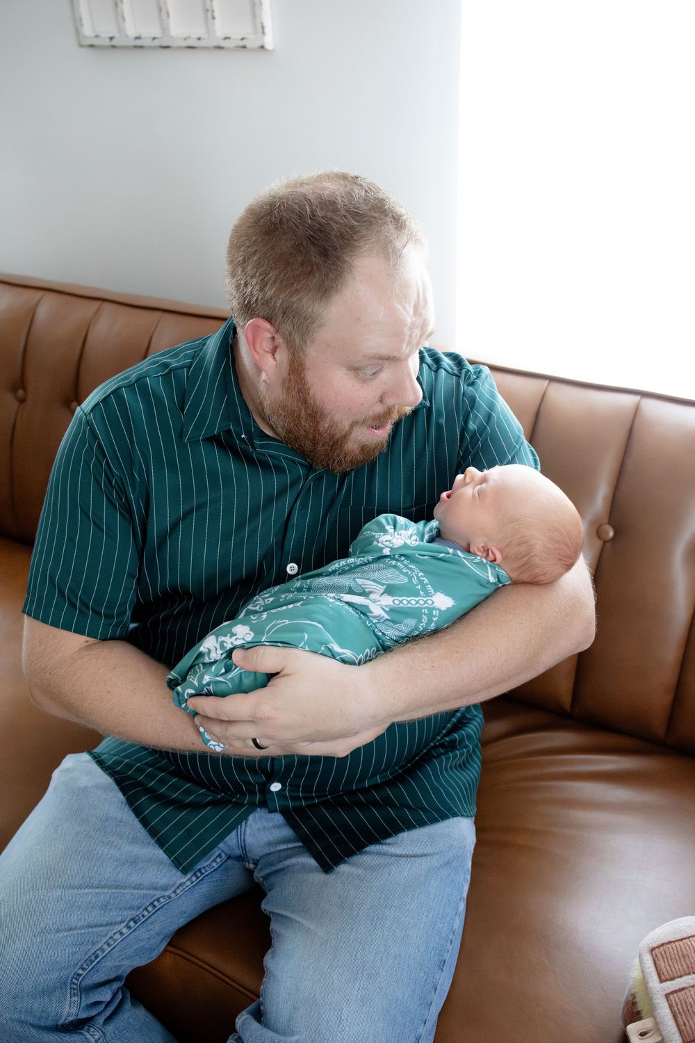 Bearded father in green shirt holds newborn baby on brown leather couch, gazing lovingly at his infant.