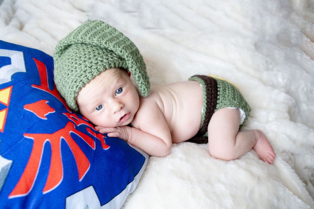 Newborn baby dressed in a crocheted green Link hat and diaper cover, resting on a Legend of Zelda Hylian Shield pillow.