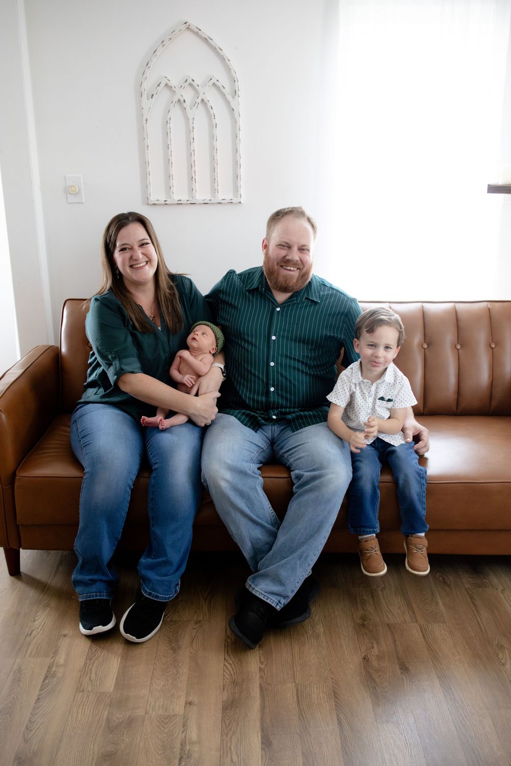 A happy family of four with a newborn and toddler poses together smiling on a brown leather couch.