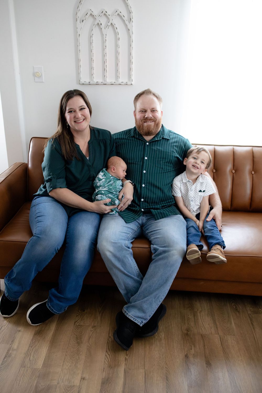 A smiling family with a newborn baby and young toddler boy sits together on a brown leather sofa.