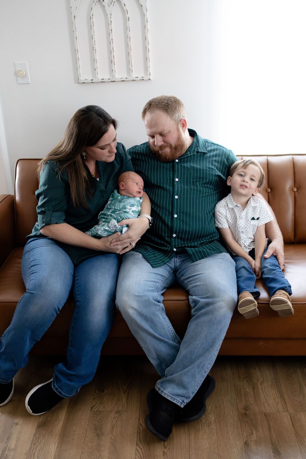 Family of four sitting on brown leather sofa, parents admiring newborn baby while toddler sits beside them.