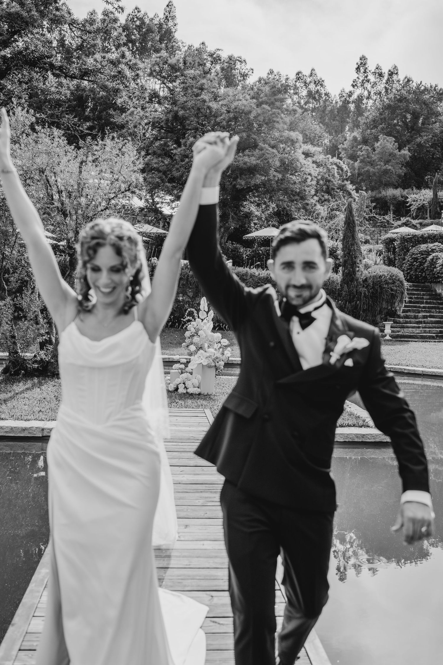 Black and white photo of a joyful bride and groom raising hands in celebration at an outdoor garden wedding venue.