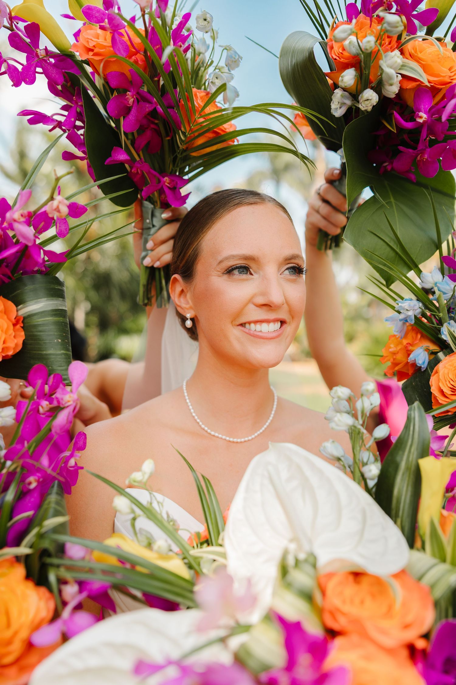 Smiling bride in white dress and veil surrounded by vibrant tropical bouquets of orange, purple, and pink flowers.