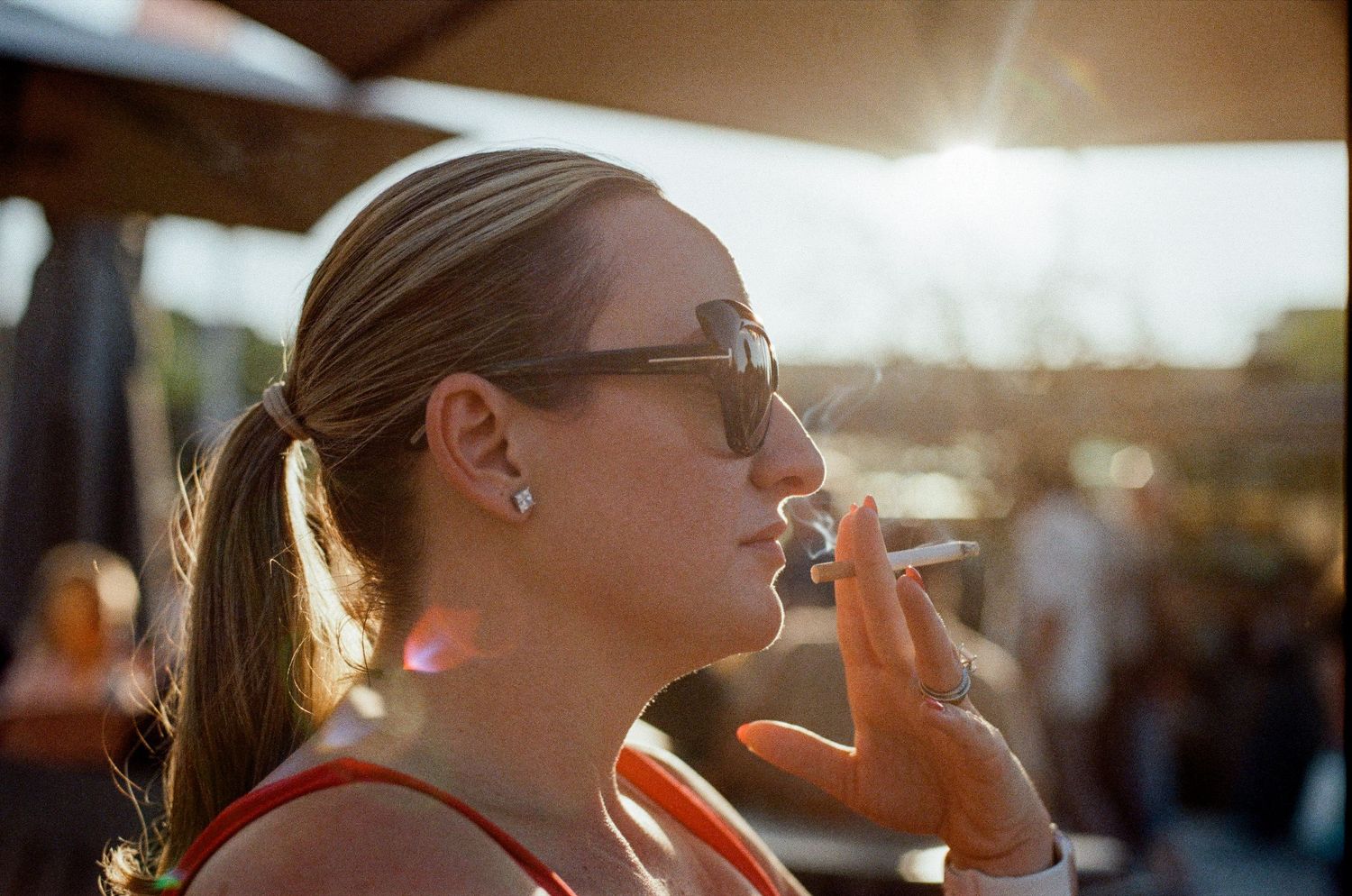 Woman in sunglasses smoking a cigarette outdoors in golden sunlight at a social gathering.