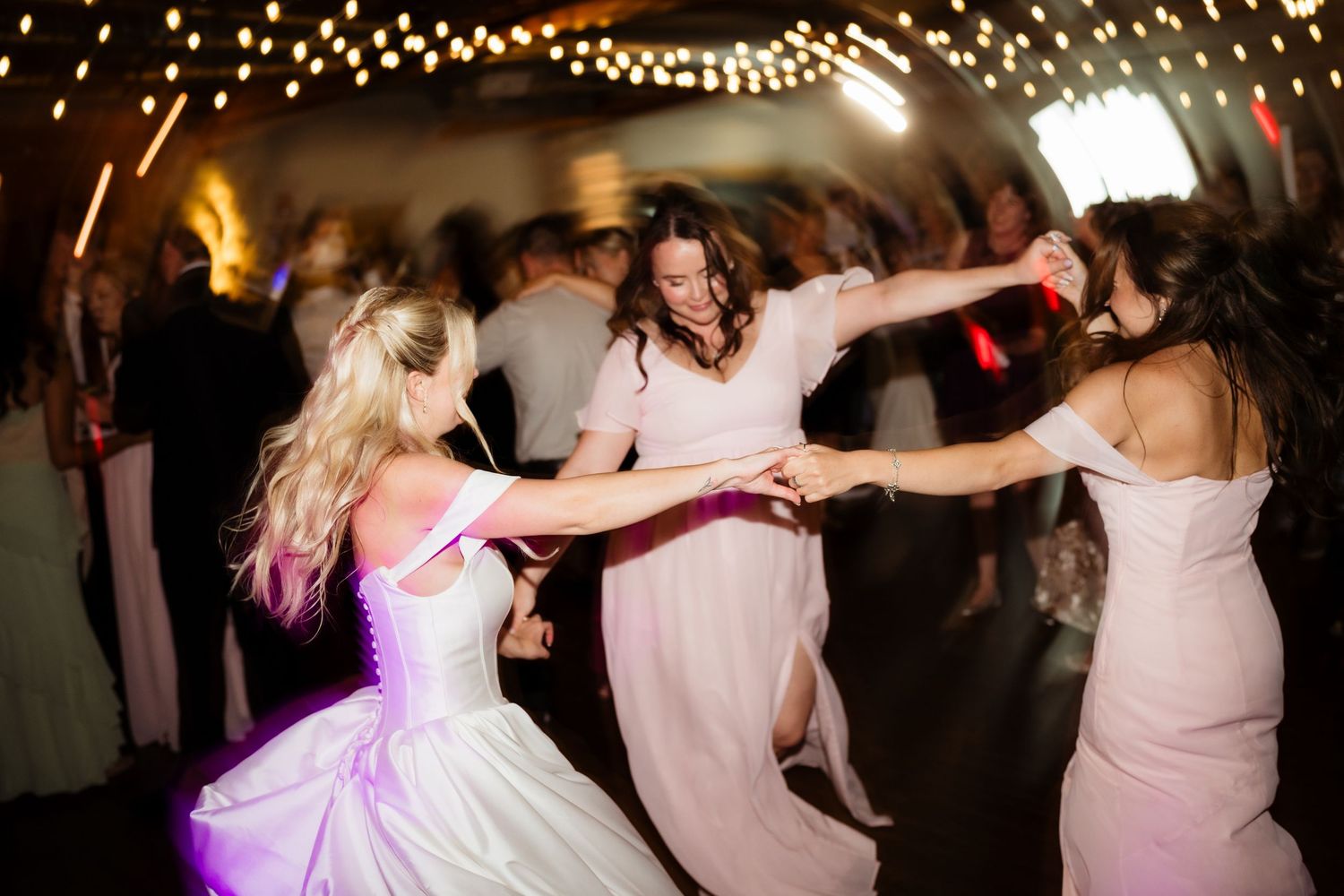 Bride and bridesmaids dancing joyfully at a wedding reception under warm string lights.