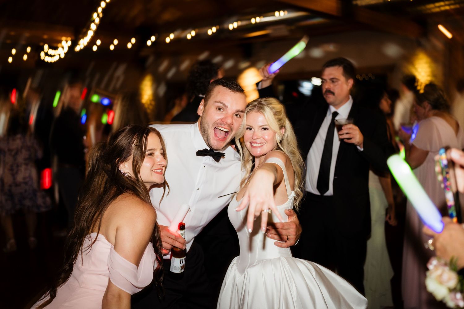 Bride and groom celebrate with guest on wedding dance floor, holding glow sticks under warm festive lights.