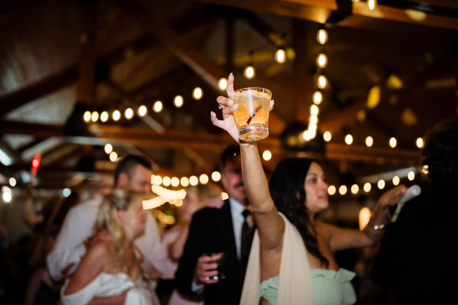 Bride raises a cocktail glass at a warmly lit wedding reception with string lights and guests dancing in background.