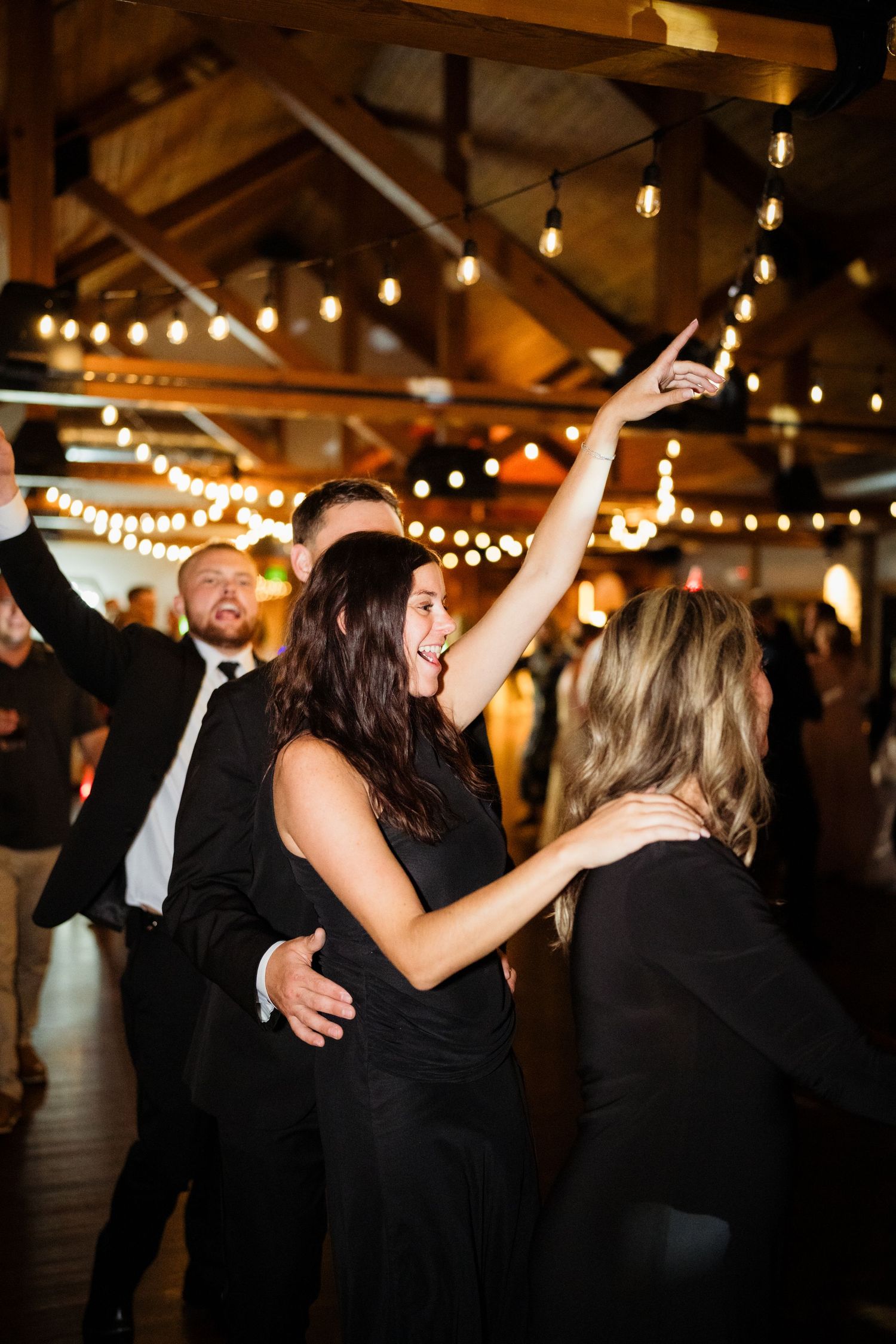 Guests dancing joyfully at a wedding reception in a rustic barn venue with warm string lights overhead.