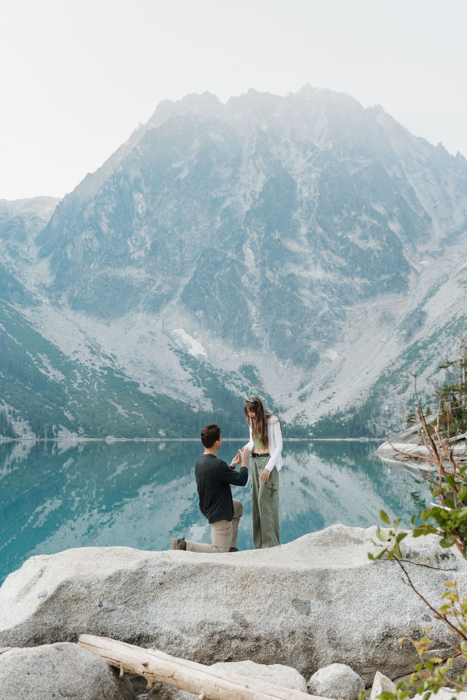 Man proposing to woman on rocky lakeside with stunning mountain reflection in turquoise alpine lake backdrop.