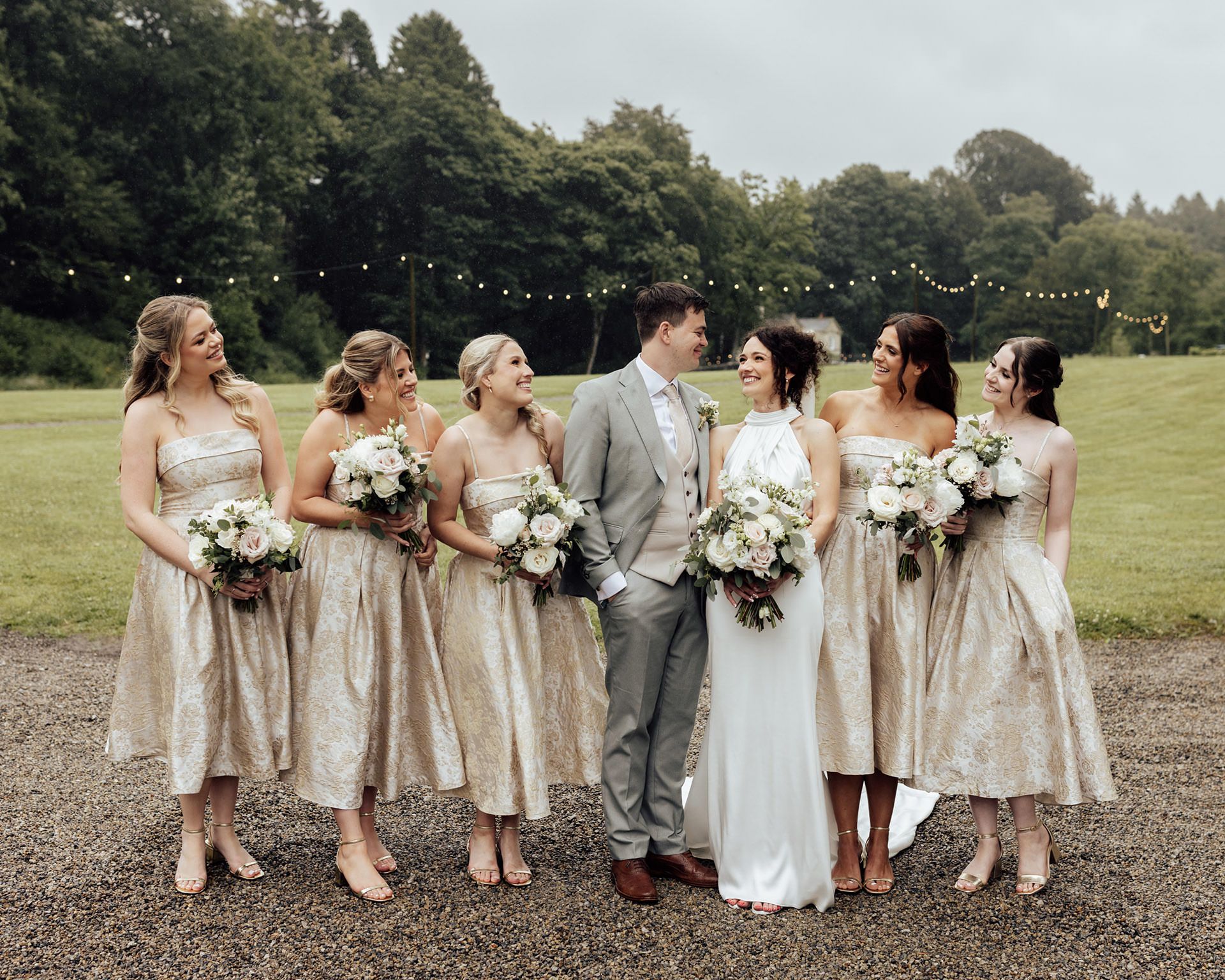 Bride in white gown with five bridesmaids in gold dresses holding white bouquets, posing outdoors at a wedding venue.