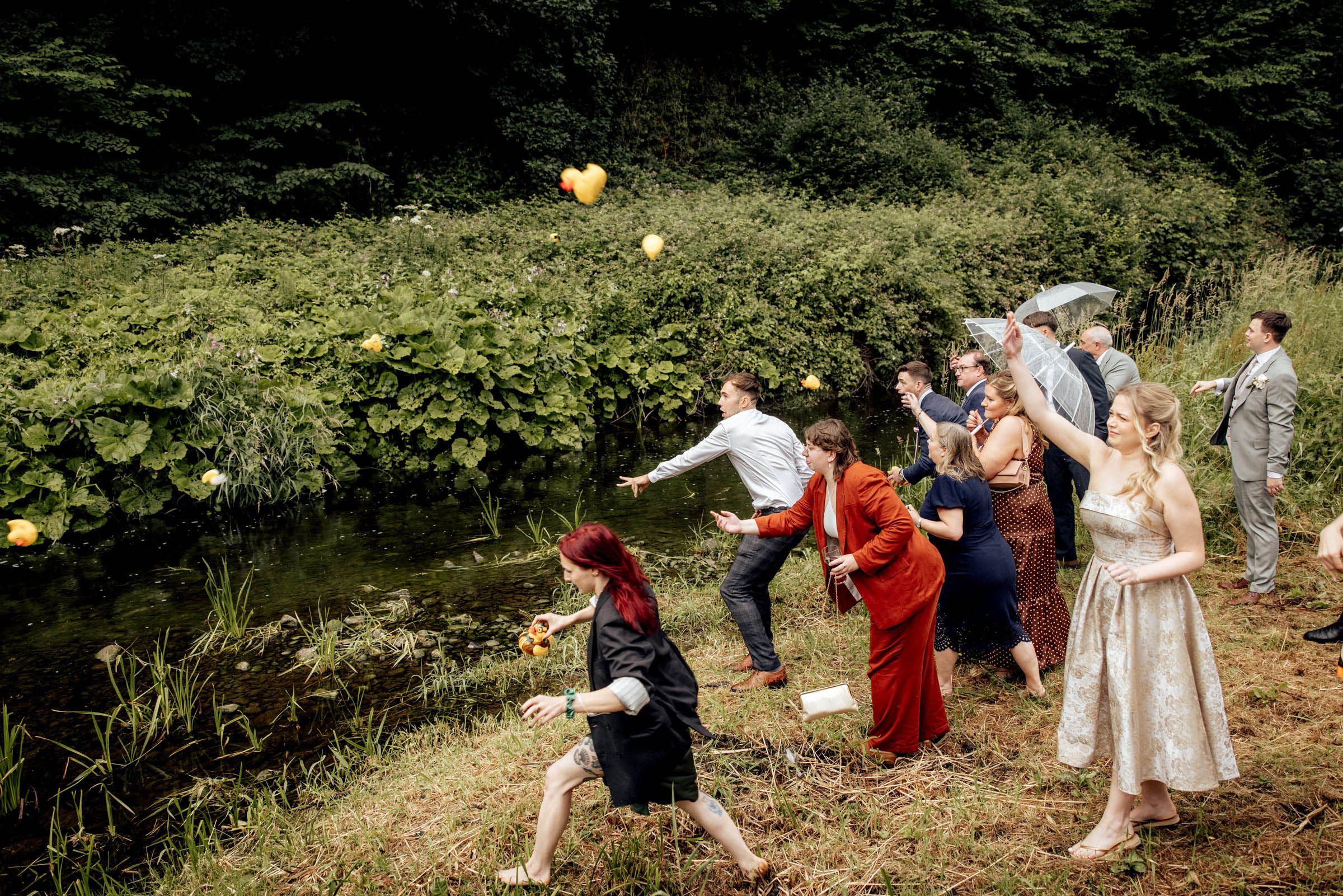 Wedding guests tossing yellow balloons near a pond surrounded by lush greenery during an outdoor celebration.