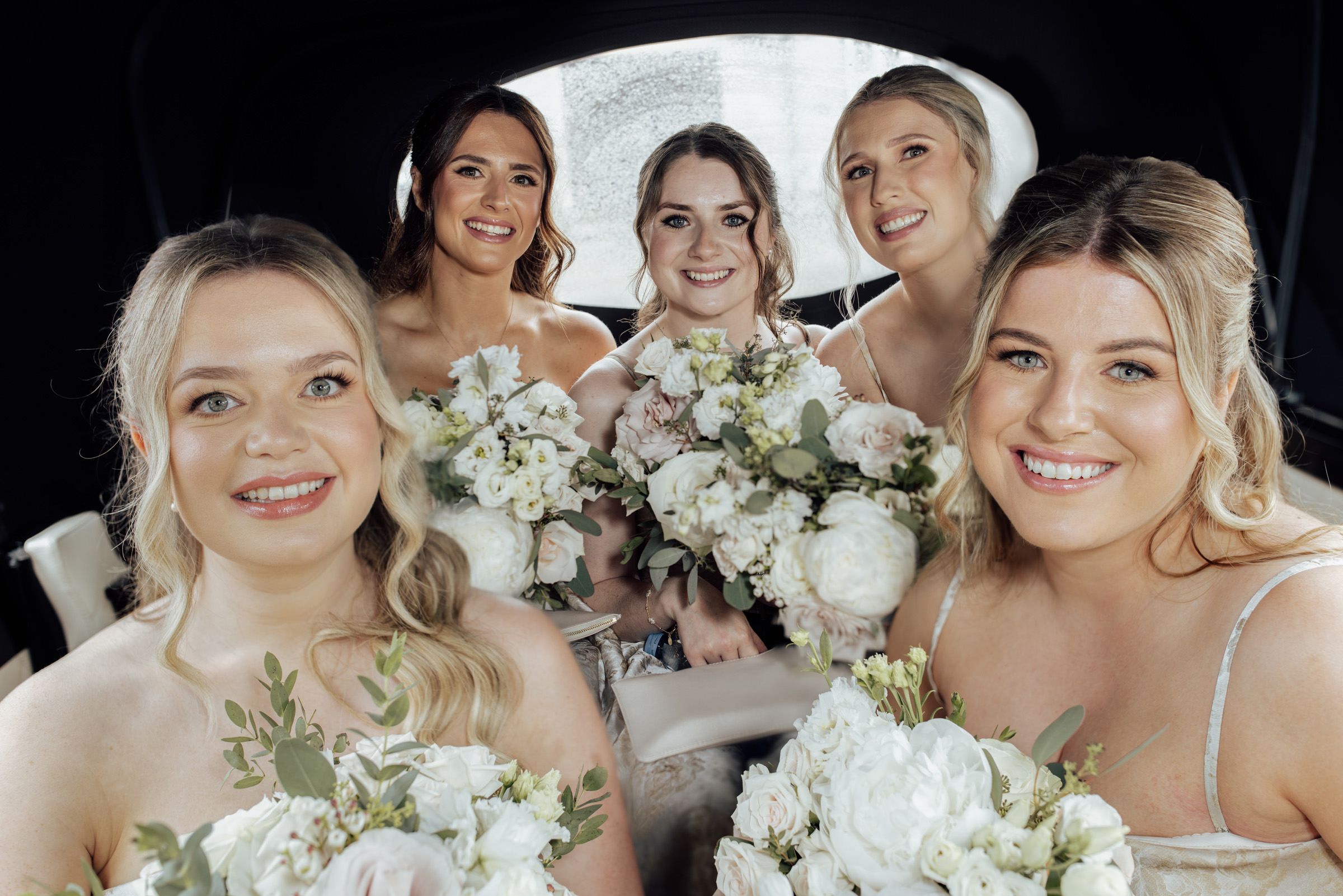 Five bridesmaids in a limousine holding white floral bouquets, smiling for a wedding photo.