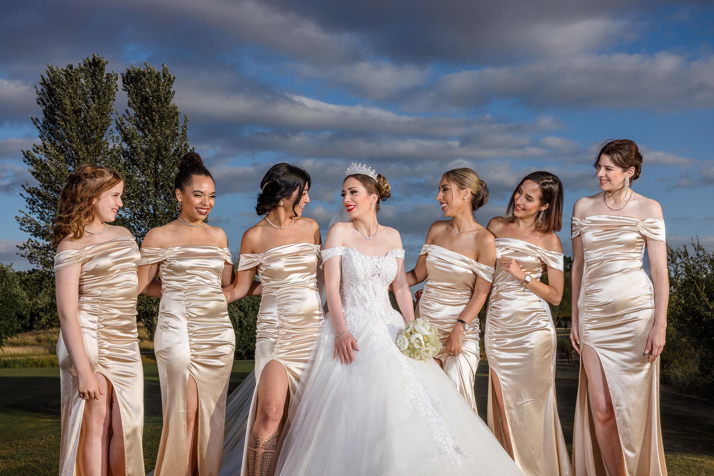 Bride in white gown with five bridesmaids in champagne satin dresses posing outdoors under a dramatic cloudy sky.