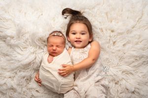 Newborn baby photo with toddler sister; newborn baby wrapped in white with toddler sister posed on fur rug looking up