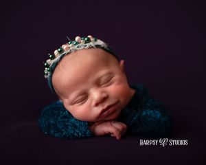 Luxury newborn photography Lexington, SC; smiling newborn baby girl wearing blue outfit & flowers & headband on purple backdrop; jewel-tones