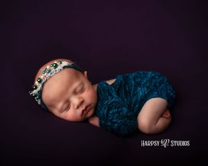 Luxury newborn photography Lexington, SC; baby girl wearing a jewel-toned blue onesie with flowers, posed on purple backdrop; newborn photo