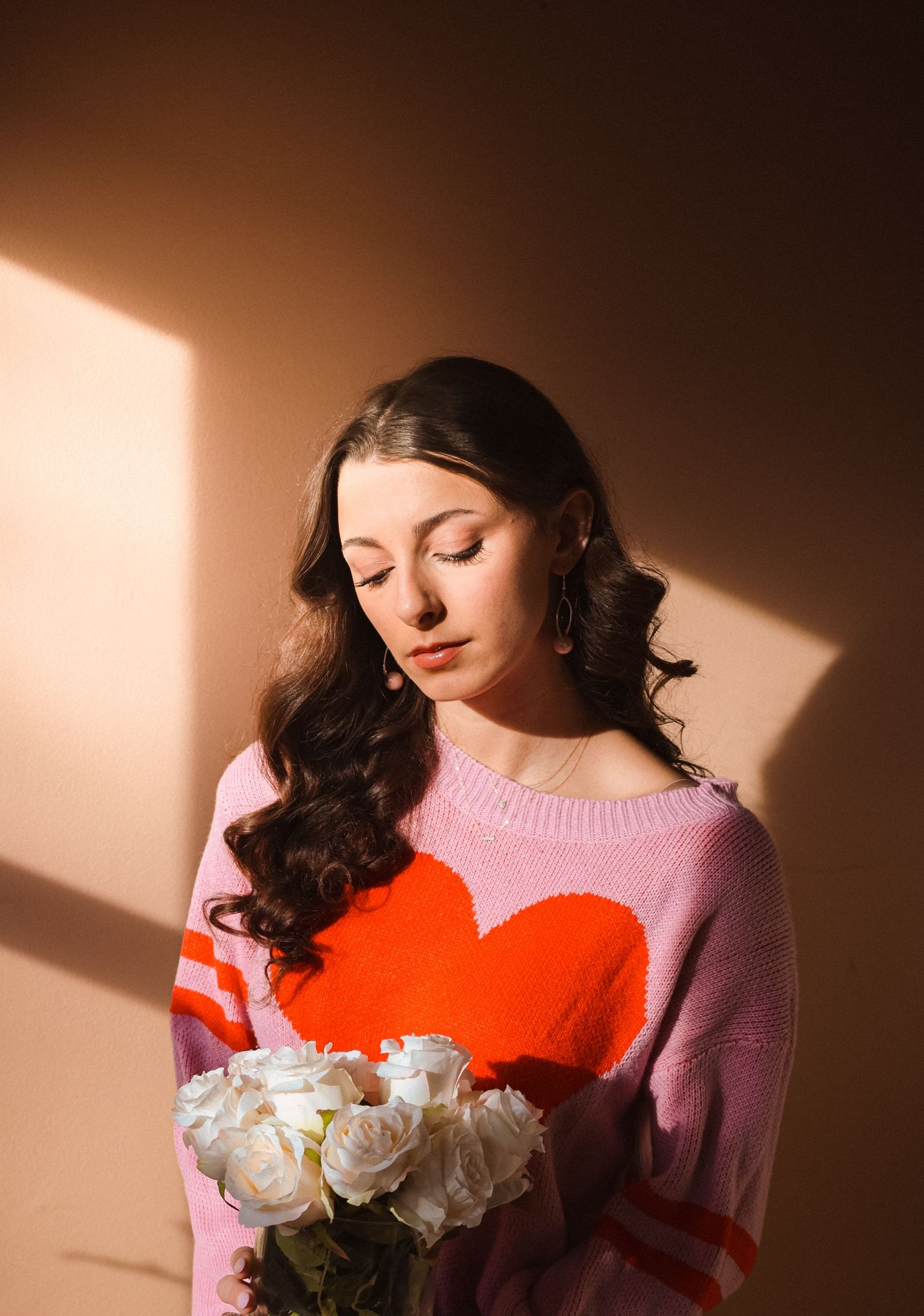 Moody portrait with pink heart sweater and white roses in dramatic lighting.