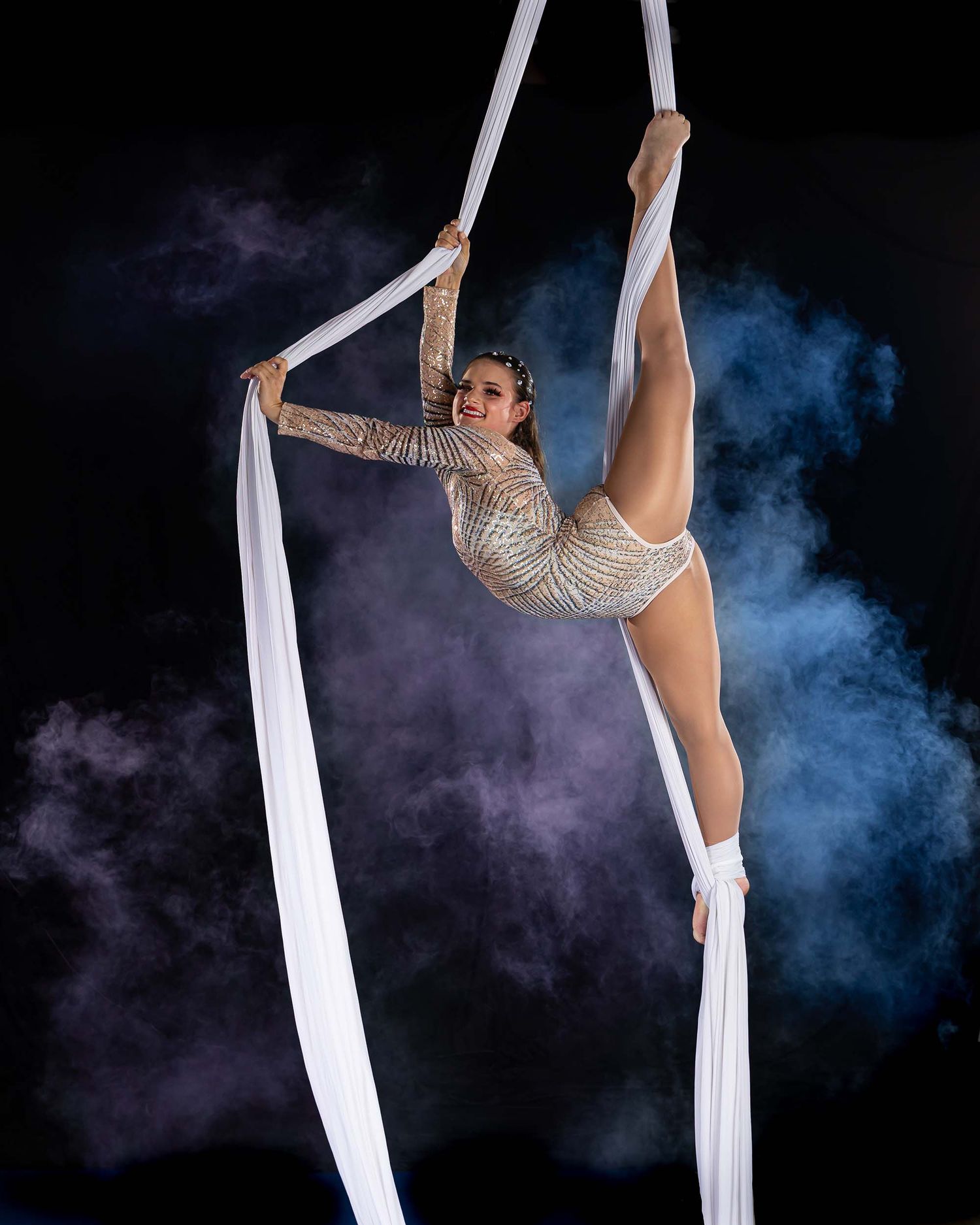 Aerial performer in sparkly costume strikes graceful pose on white silks with smoke effects.