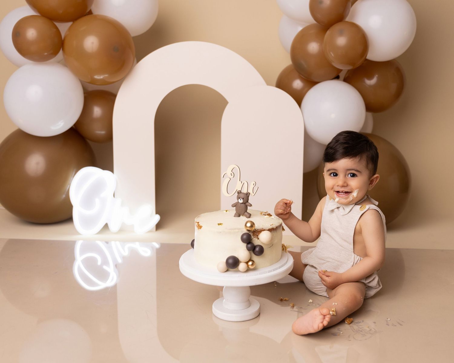 Baby's first birthday setup with brown and white balloons, an arch, and cake on white pedestal.