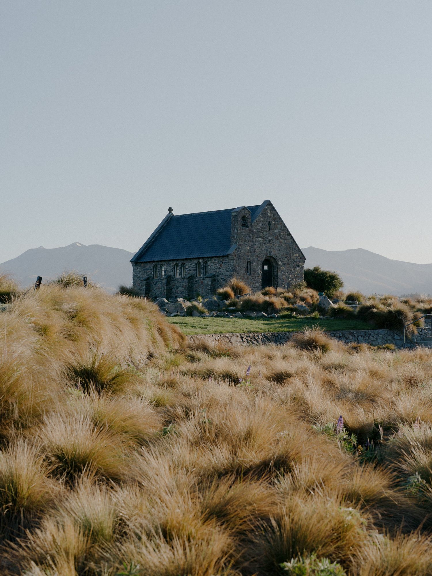 Stone church building stands alone on grassy hillside with tussock grass and mountains in background.