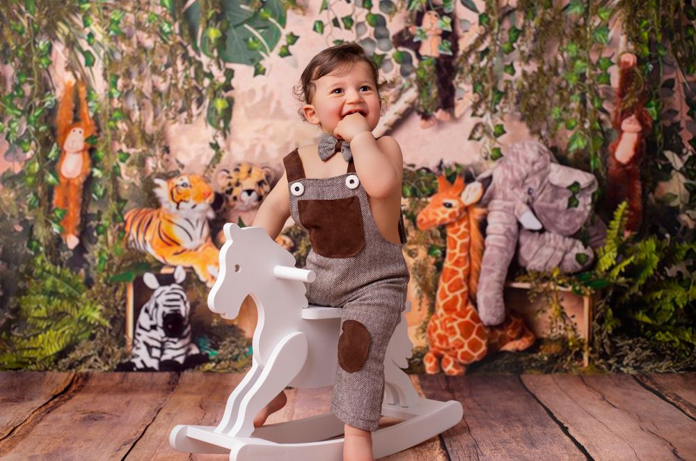 Baby wearing grey romper sits on white wooden rocking horse toy against floral backdrop.