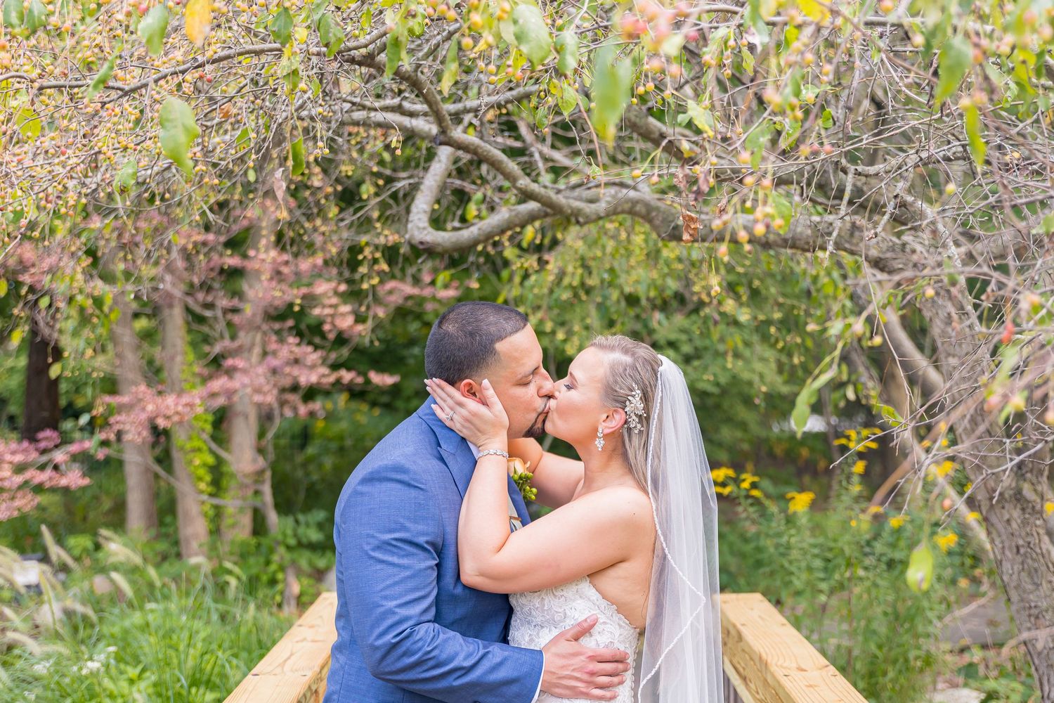 A romantic wedding couple shares an intimate kiss on a wooden bridge surrounded by autumn foliage.