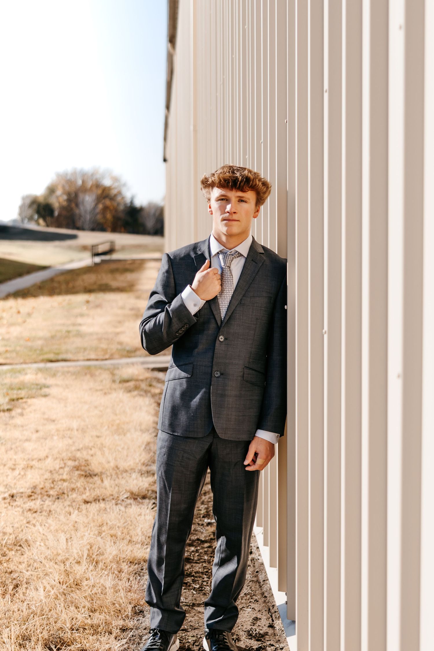 Figure in grey suit poses thoughtfully against white architectural columns outdoors.