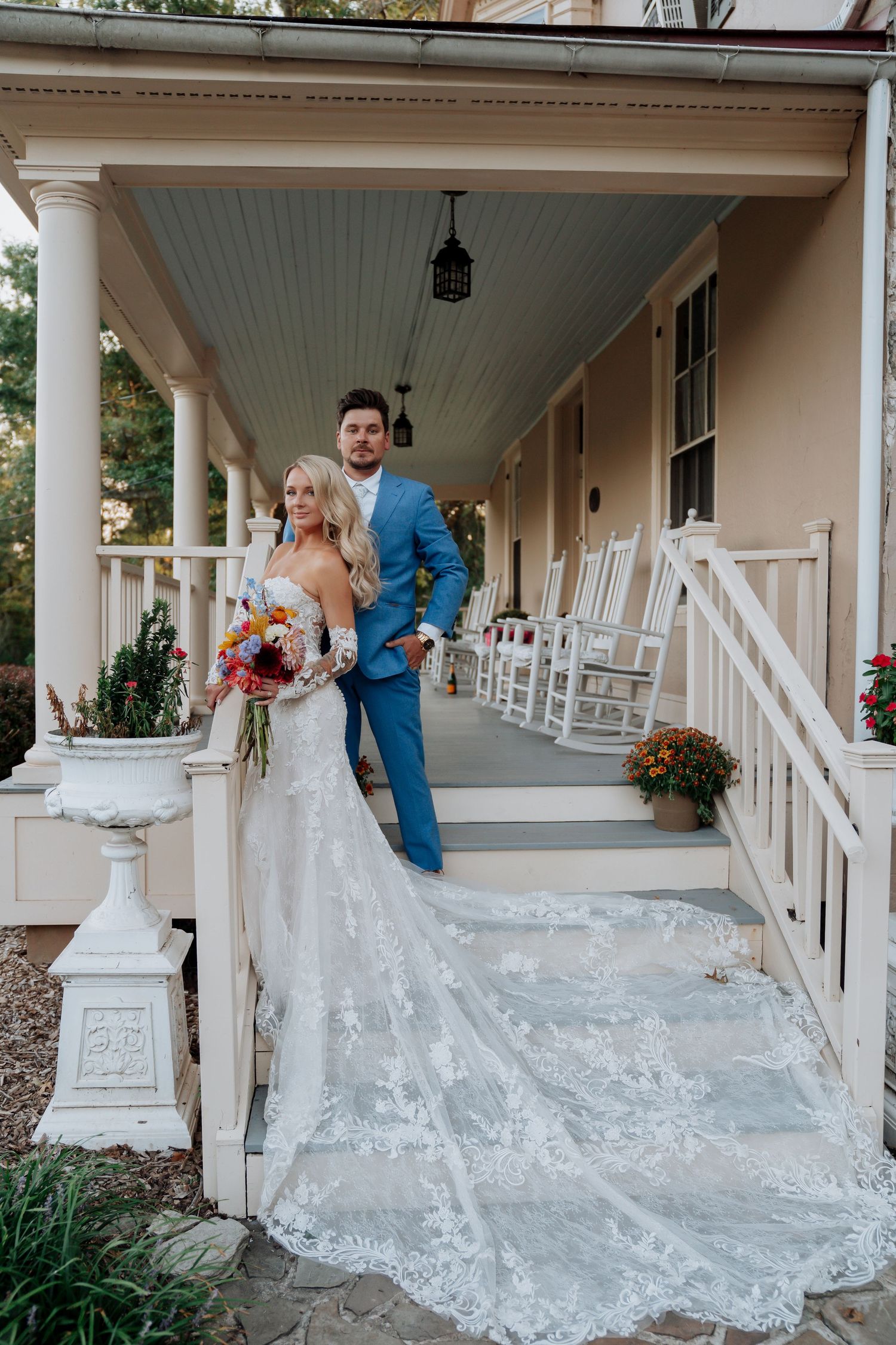 Bride and groom pose on a charming Southern porch with white rocking chairs and a lace wedding gown train.