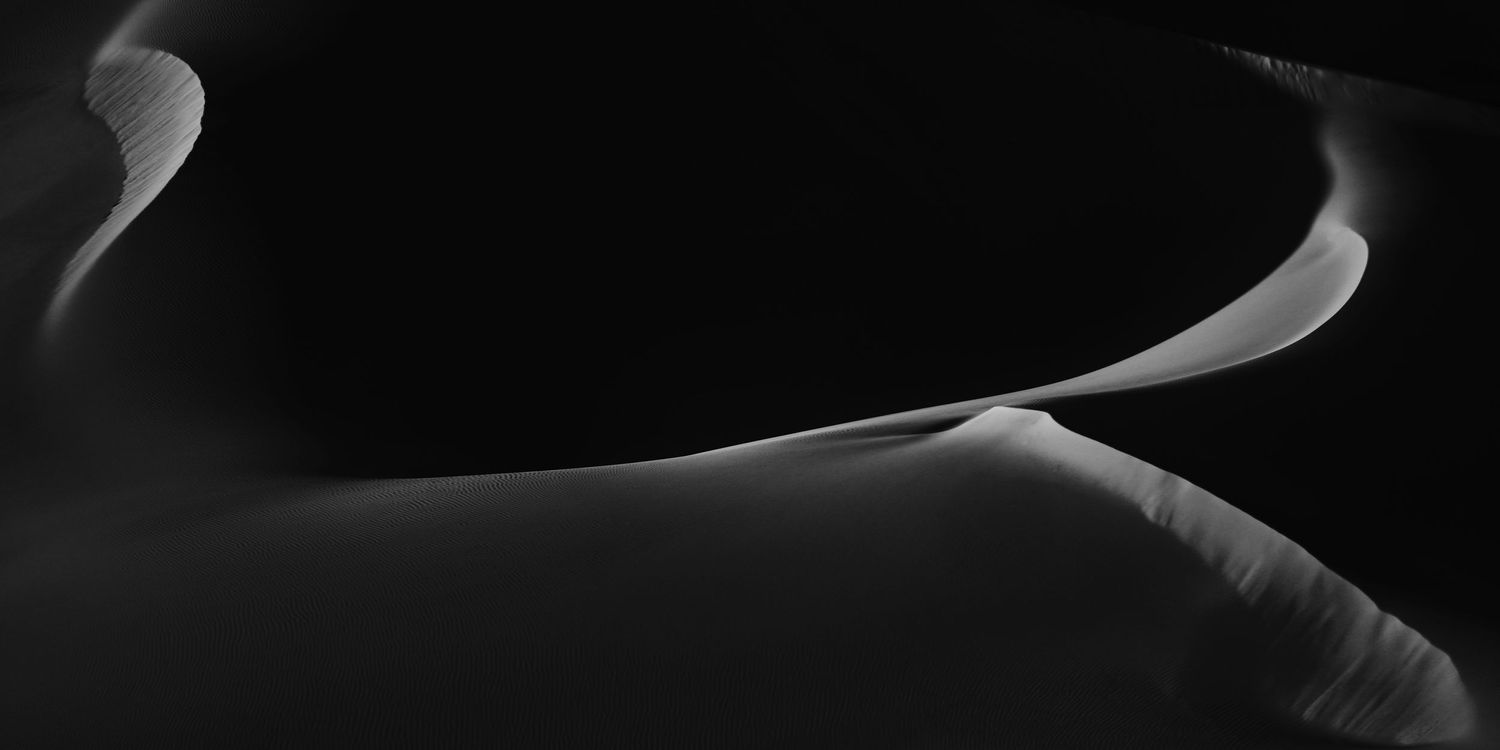 Black and white abstract desert sand dune landscape with smooth curves and shadows creating dramatic contrast.