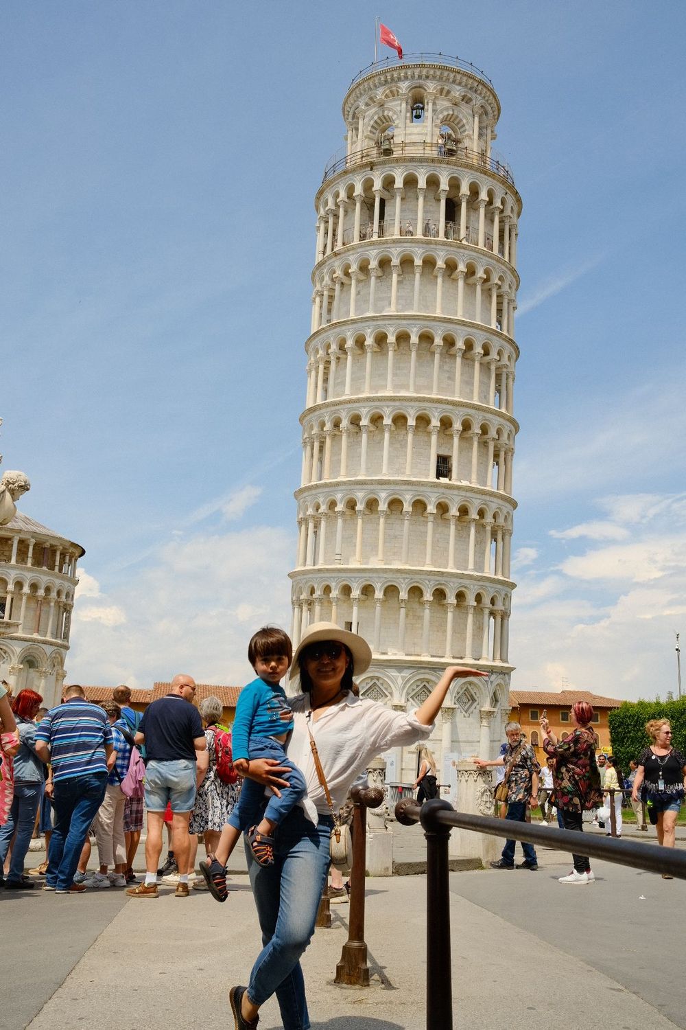 Tourist poses near the iconic Leaning Tower of Pisa in Italy on a sunny day surrounded by other visitors.