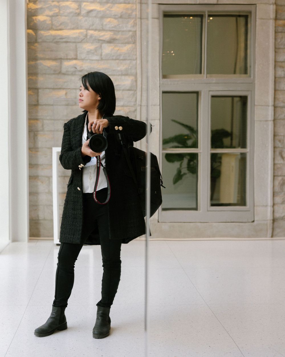 Stylish woman in black outfit stands against light colored wall next to window.