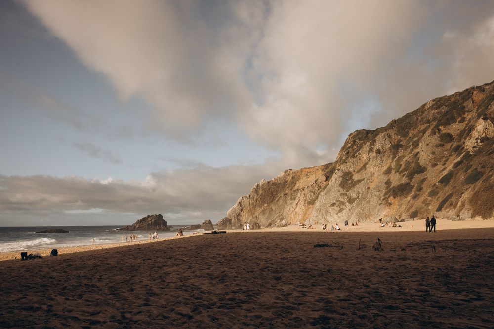 Dramatic coastal cliffs and sandy beach stretch along the shoreline under a cloudy sky at sunset.