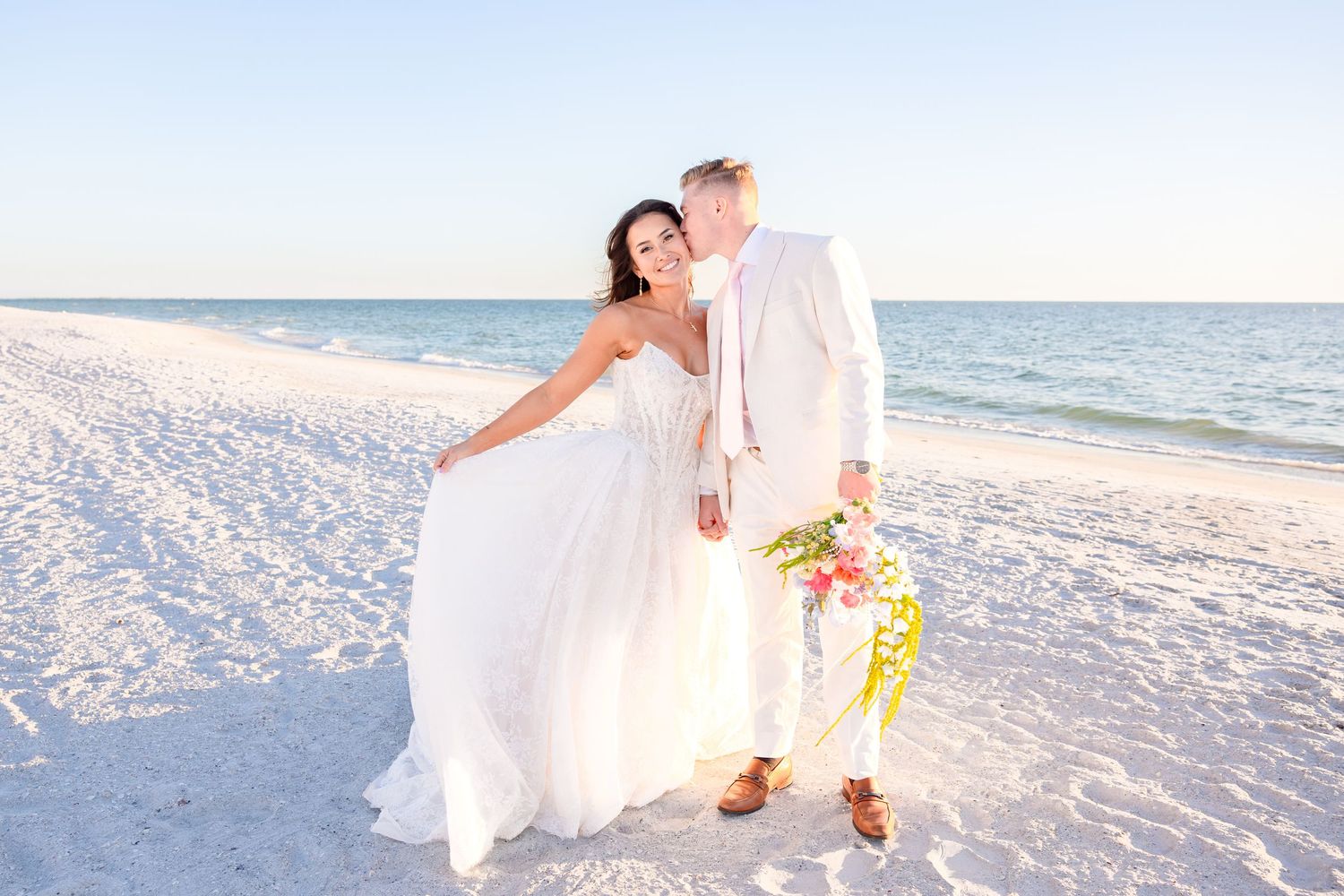 Bride in white long-sleeve gown embraces groom in black tuxedo outdoors among lush greenery.