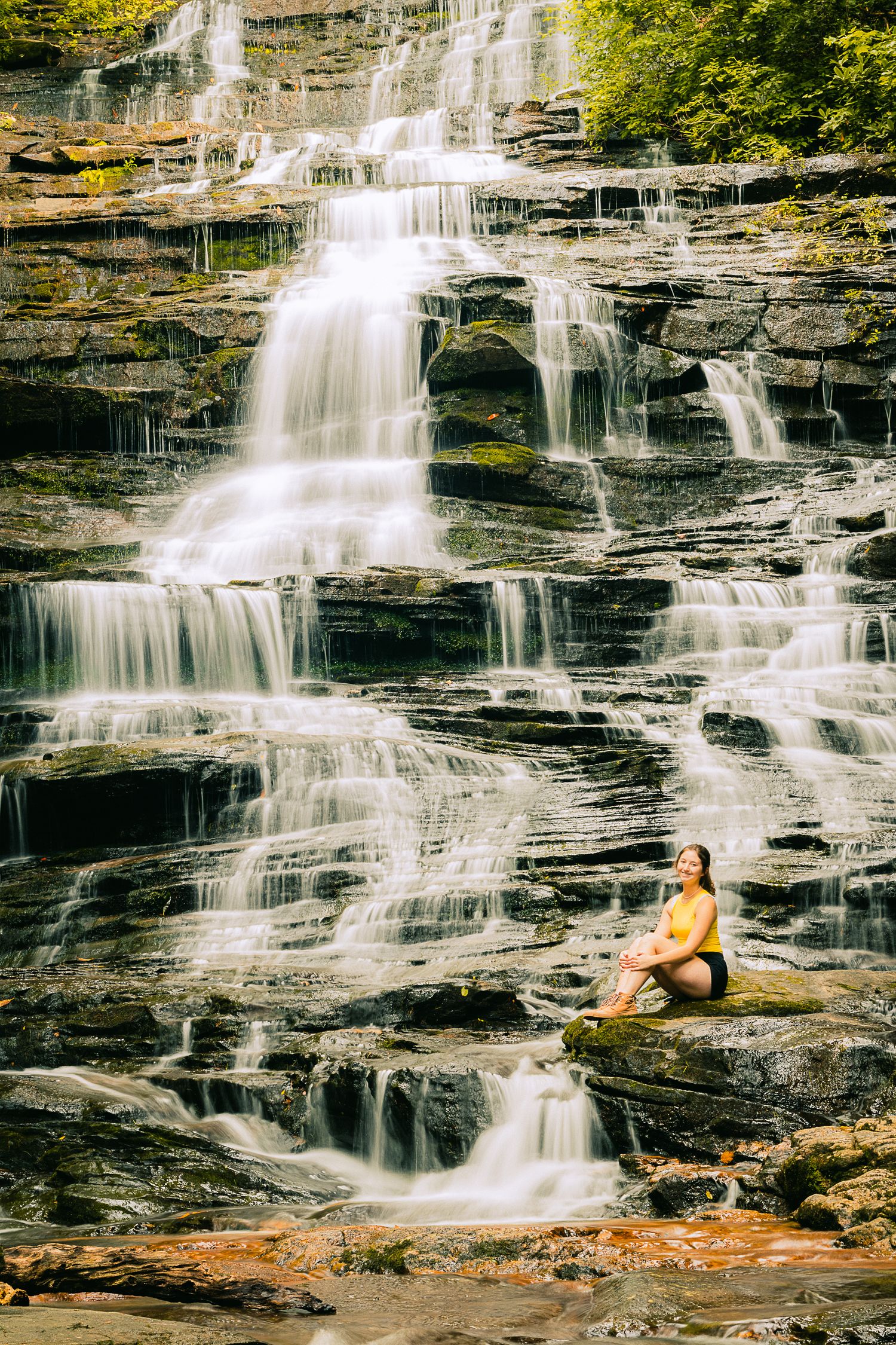 A person in yellow sits on cascading waterfalls surrounded by rocky cliffs.