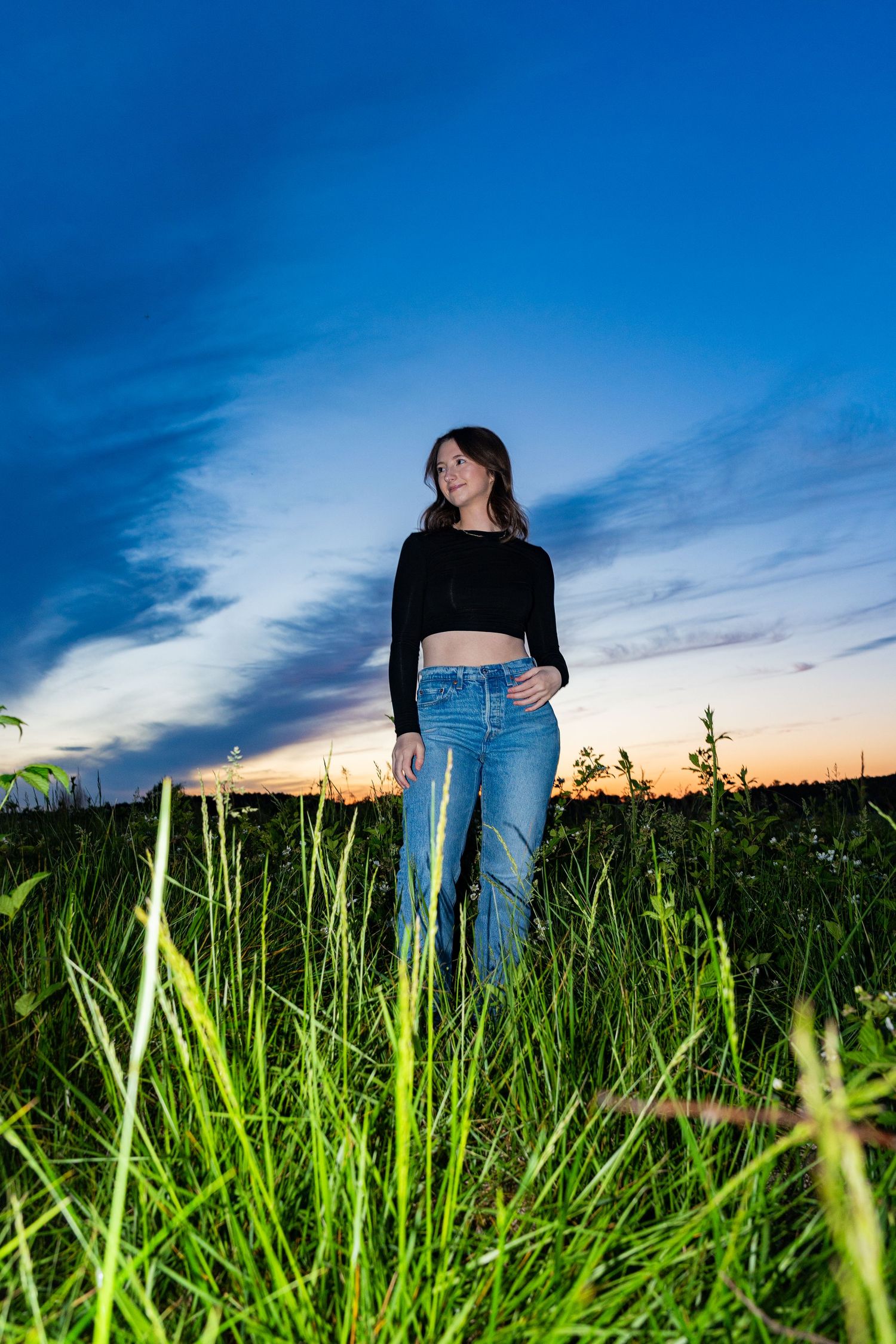 A couple stands in tall green grass against a vibrant blue sky at sunset, creating a romantic silhouette effect.