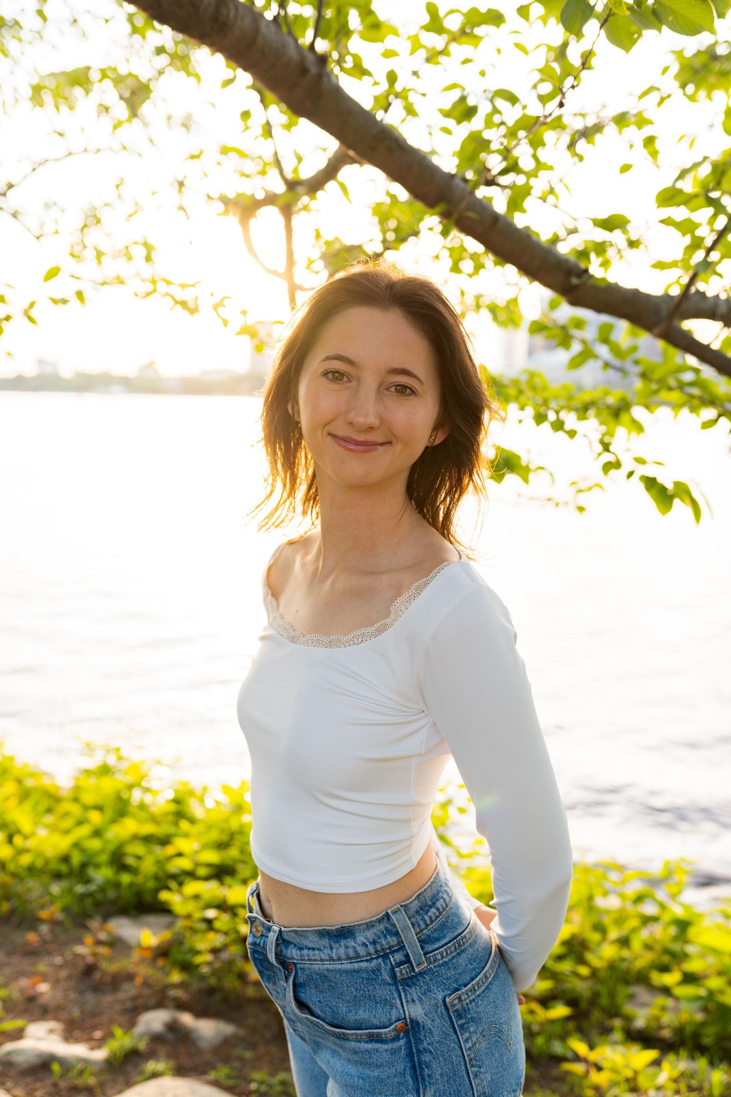 A person wearing a white long-sleeve crop top and jeans poses outdoors during golden hour under tree branches.
