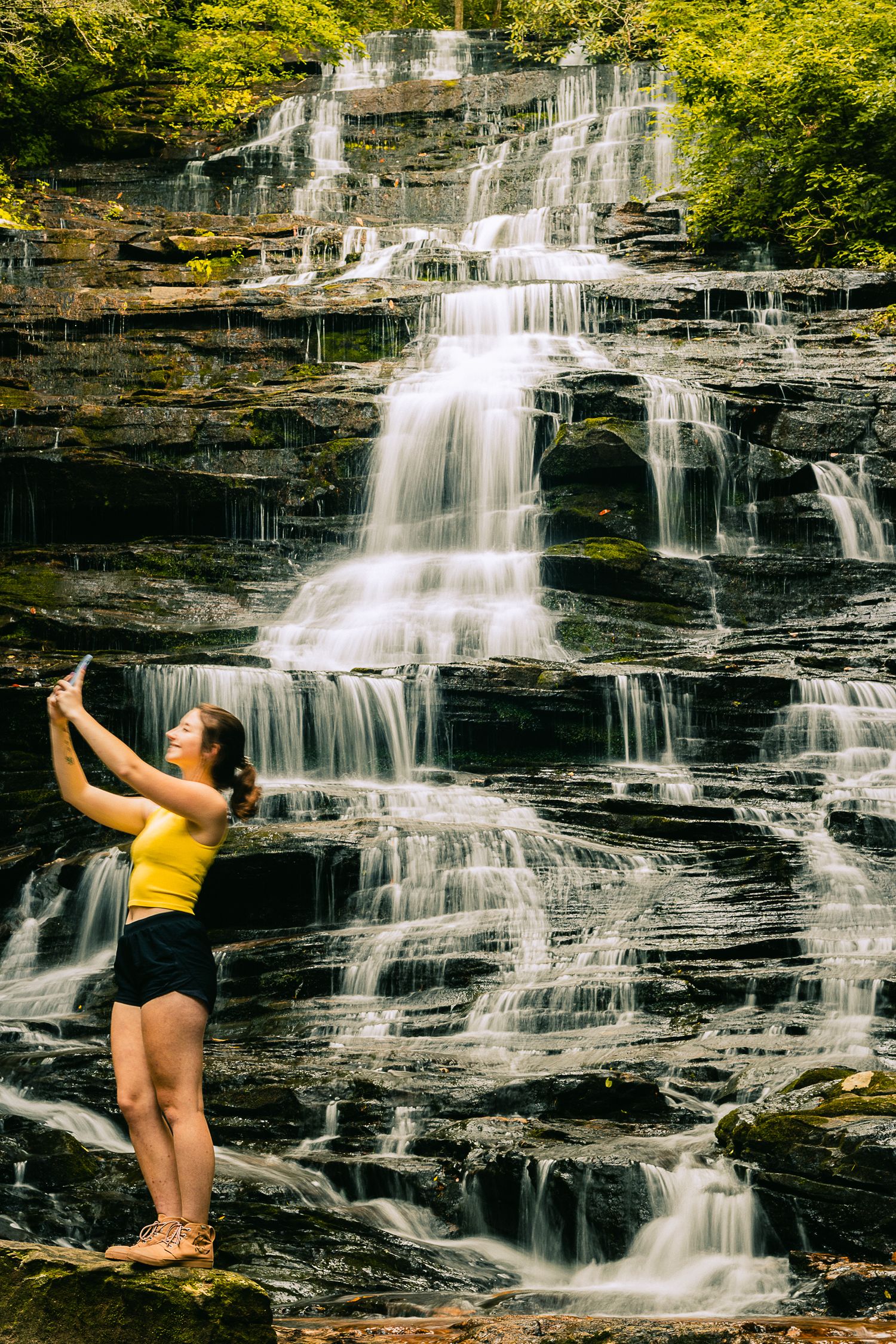 A person stretches against a majestic multi-tiered waterfall in a forest setting.
