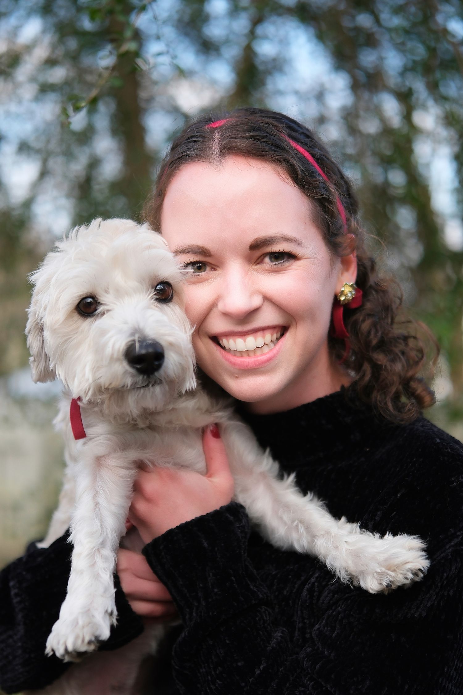 Person in black sweater holding small white fluffy dog outdoors.