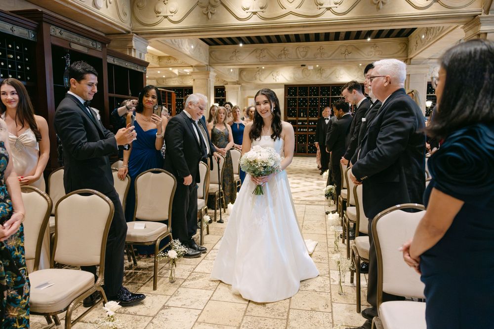 A bride in a white wedding dress walks down an elegant indoor aisle lined with cream-colored chairs during a ceremony.