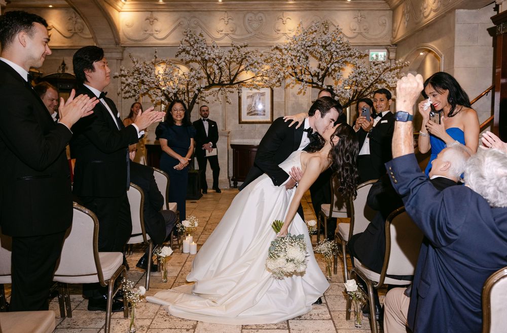 A romantic dip kiss between a couple at an elegant indoor wedding ceremony while guests look on and celebrate.