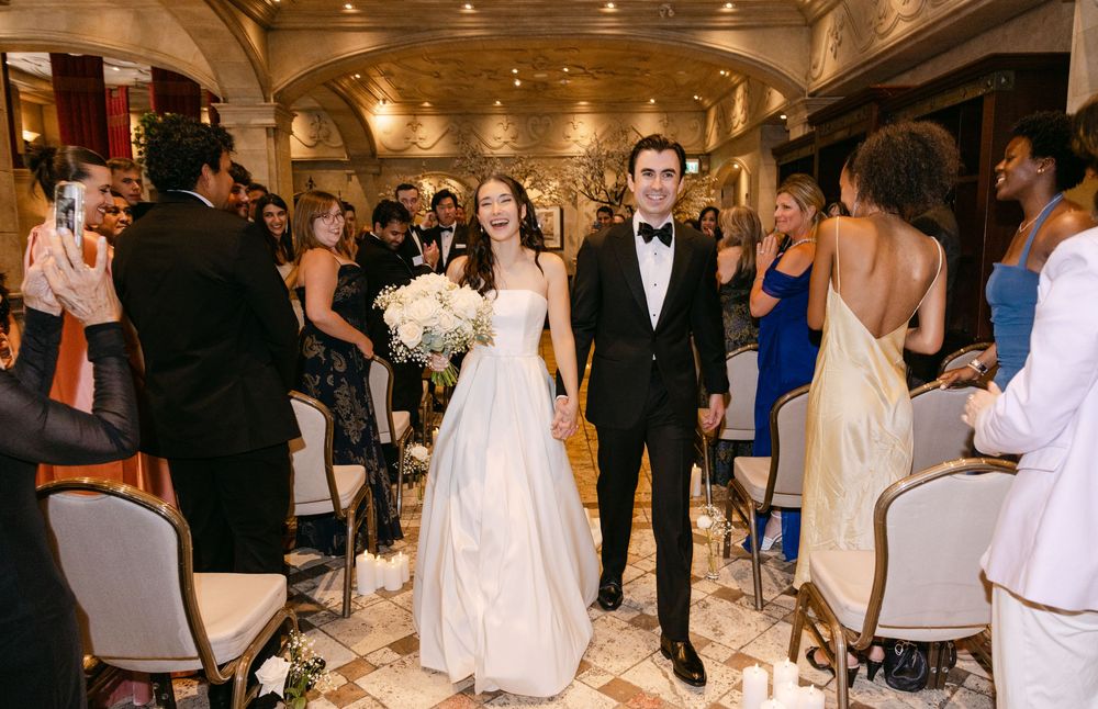 A newlywed couple walks down a luxurious hotel aisle lined with guests in elegant formal attire.