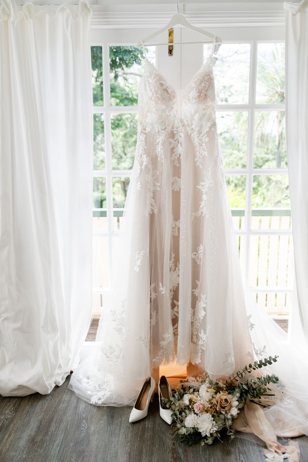 Elegant bridal gowns hang in a sunlit room with white curtains and floral arrangements on the floor.