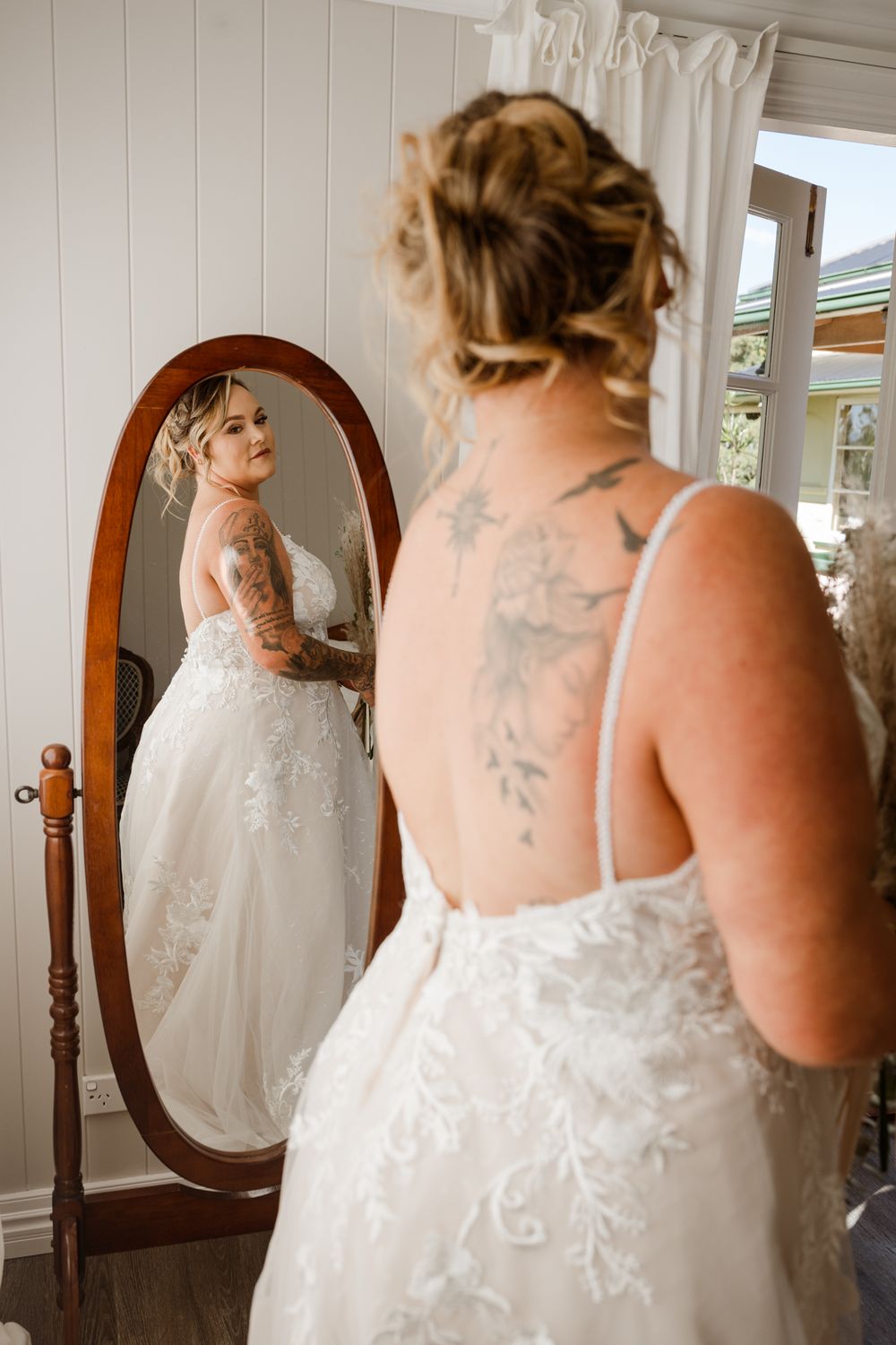 A person in a white lace wedding dress views their reflection in a traditional wooden-framed mirror.
