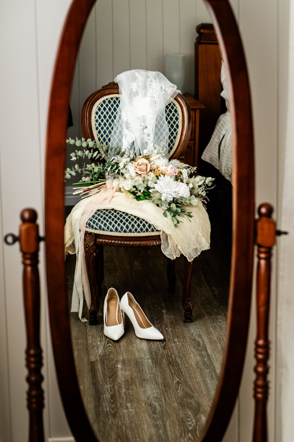 White bridal heels and floral bouquet arranged on a vintage wooden chair with an oval mirror in the background.