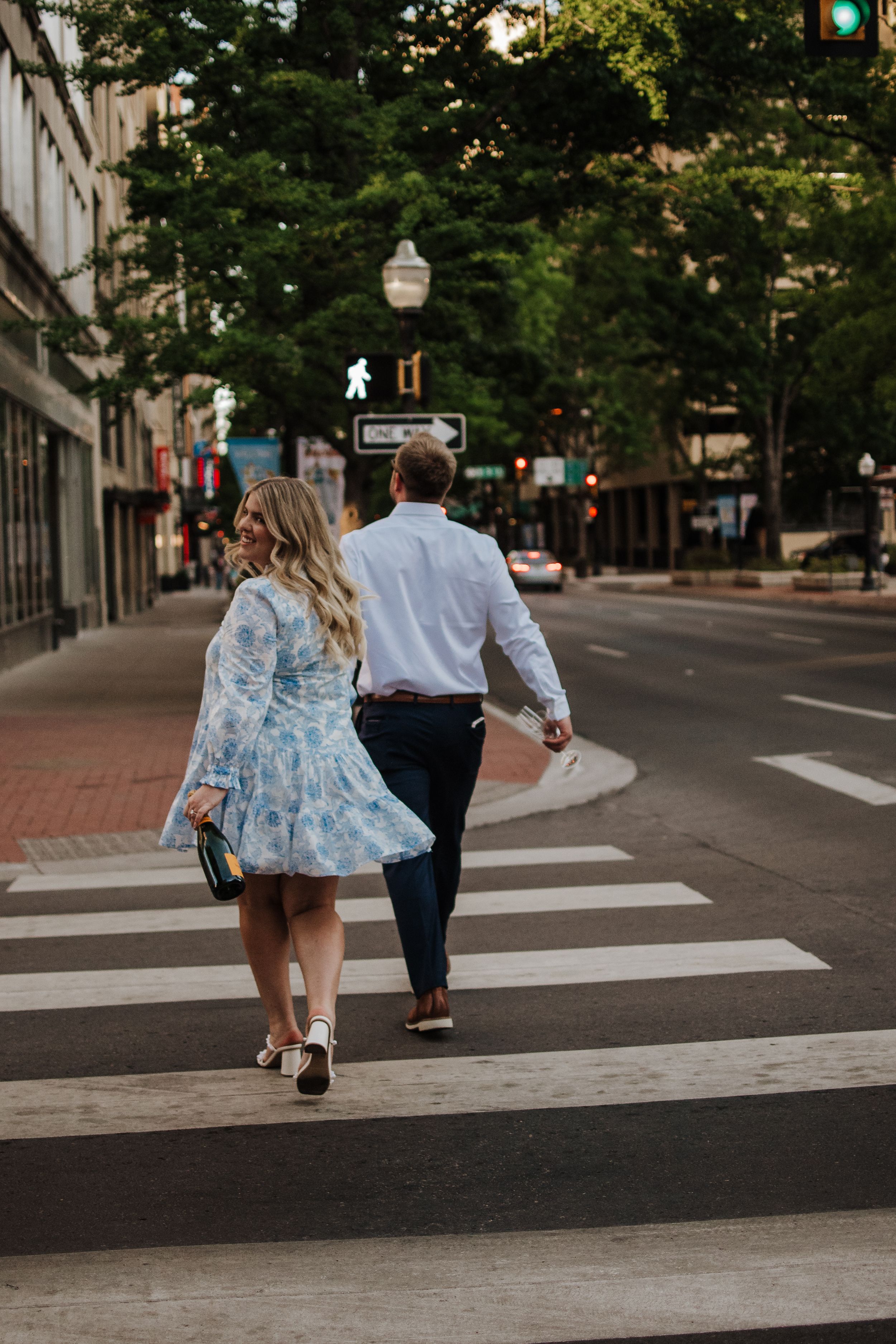 A couple walks hand in hand across a crosswalk on an urban street lined with tall buildings and trees.