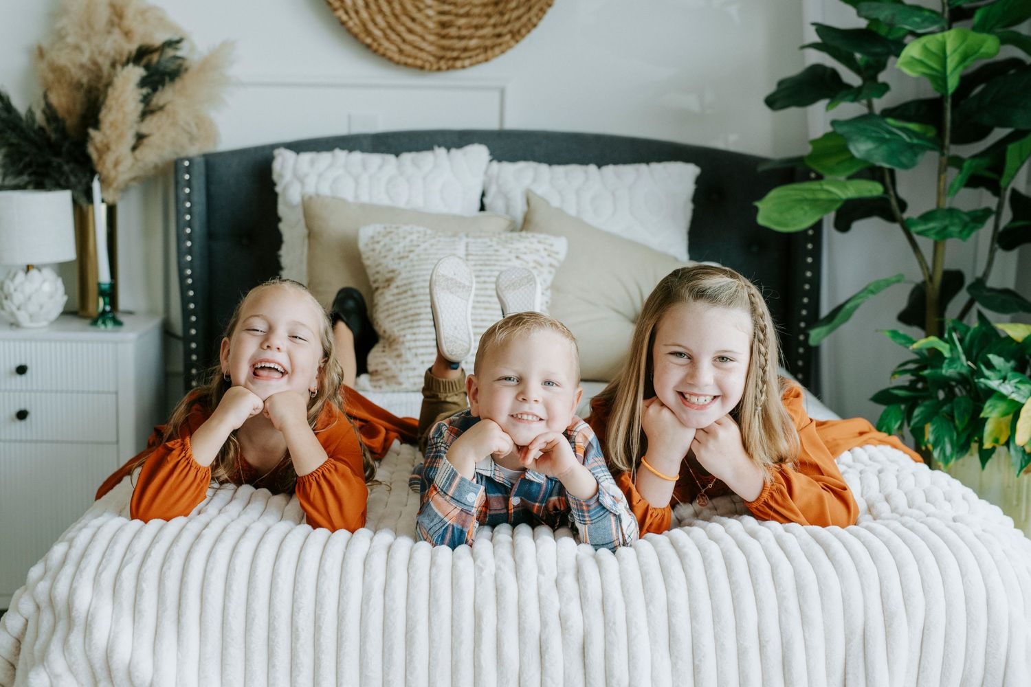 Happy kids in matching orange hooded sweatshirts lay playfully on a couch with houseplants in the background.