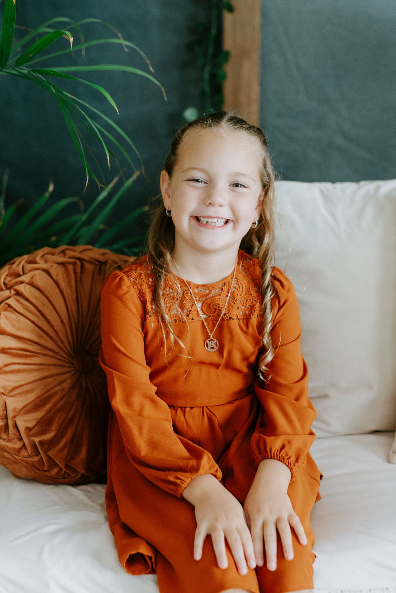 A child wearing an orange long-sleeve dress sits on a white couch against a green plant background.