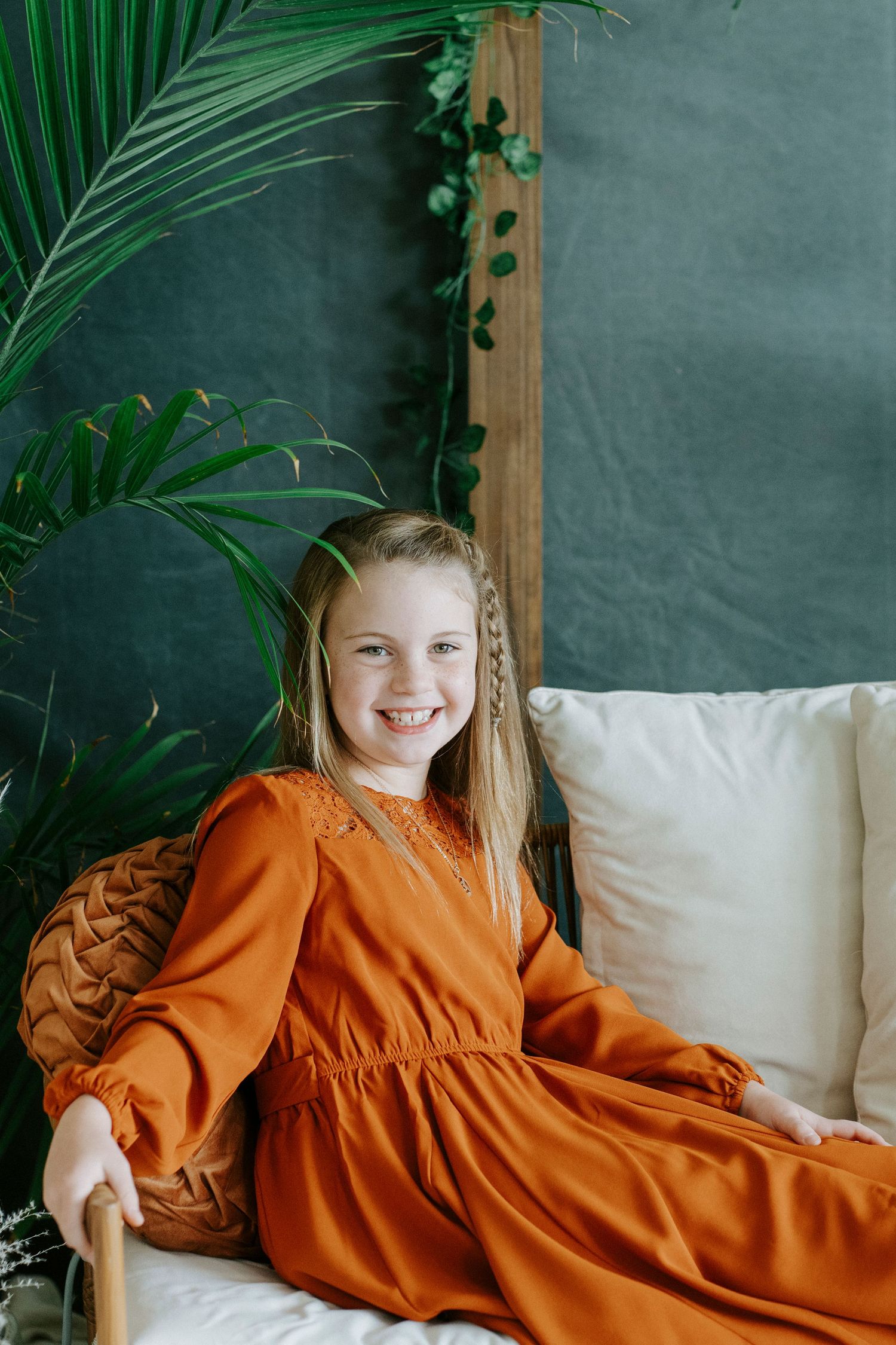 A figure wearing a rust-colored dress lounges comfortably on a white couch with tropical plants in the background.